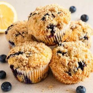 Golden vegan blueberry muffins with crumble topping on a white-beige counter, one cut open showing fluffy interior with juicy blueberries